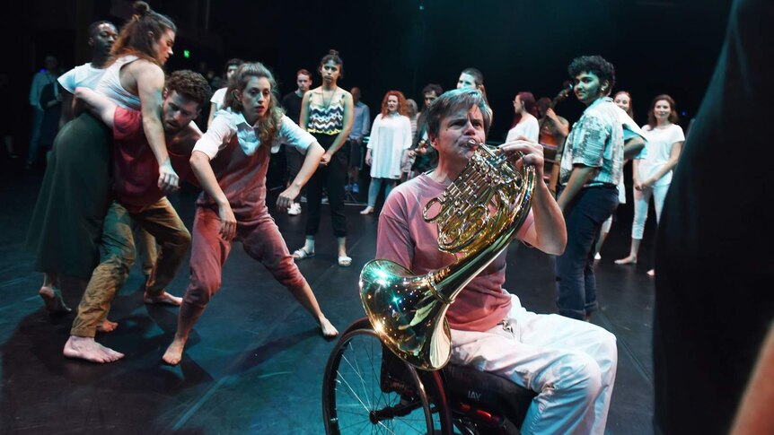 Group of people on stage, man in wheelchair playing French Horn in foreground, cluster of dancers to left. Audience behind.