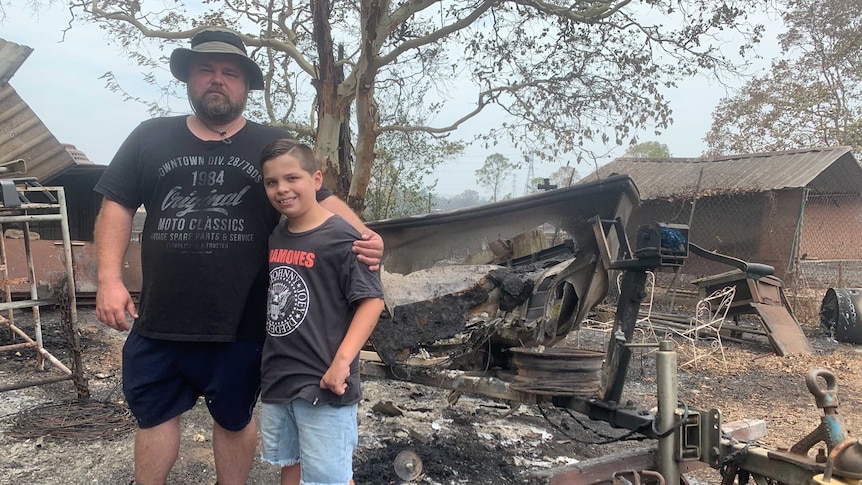 A man and a boy stand in front of a blackened, destroyed boat and on ground which is charred.