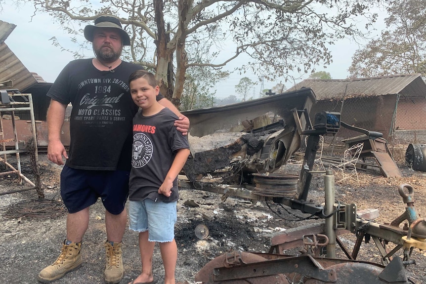 A man and a boy stand in front of a blackened, destroyed boat and on ground which is charred.