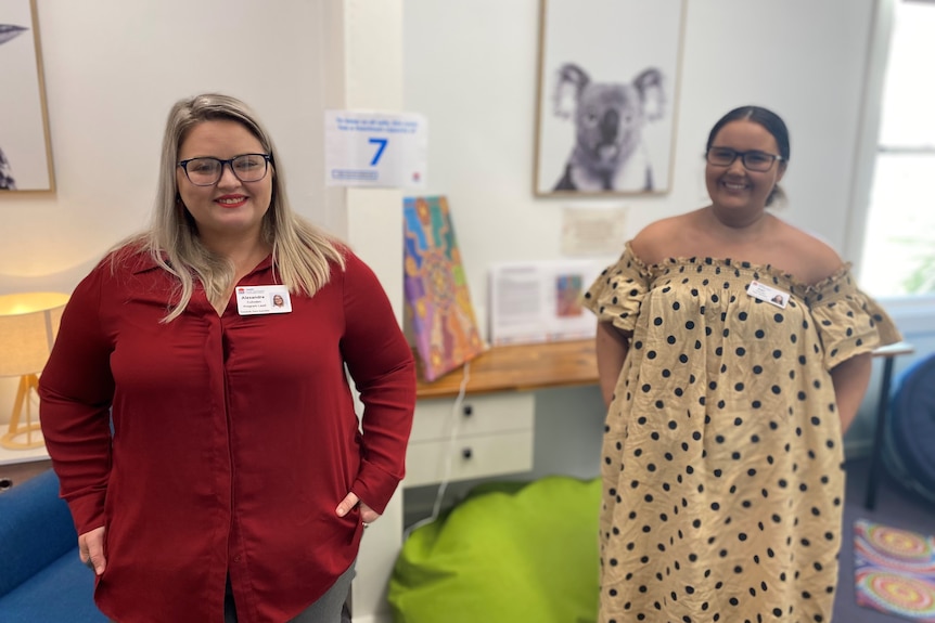 Two women, one in a red shirt, the other in a black spotted dress, stand in a colourful room. 