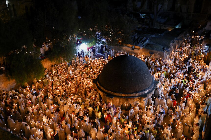 Crowds of people stand together holding candles around a domed building.