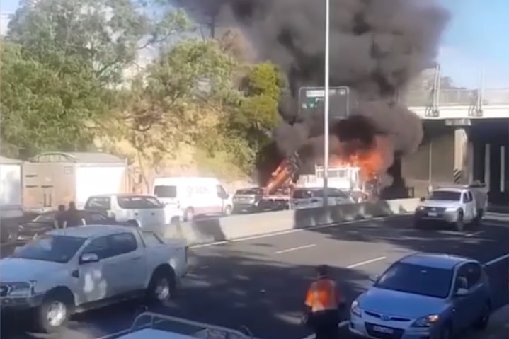 A vehicle on fire beneath an overpass on a busy motorway.