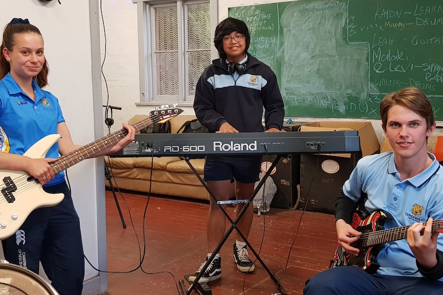 Three teenagers with guitars and a keyboard smile at the camera.