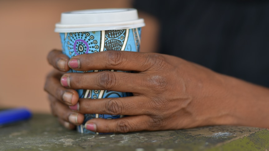 A woman's hands hold a coffee cup which has bright patterns on it. A lighter is also on the table.