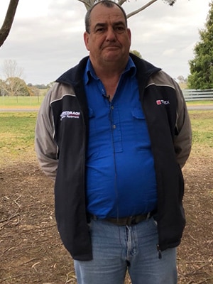 Man stands under tree wearing blue button up top and open grey jacket.