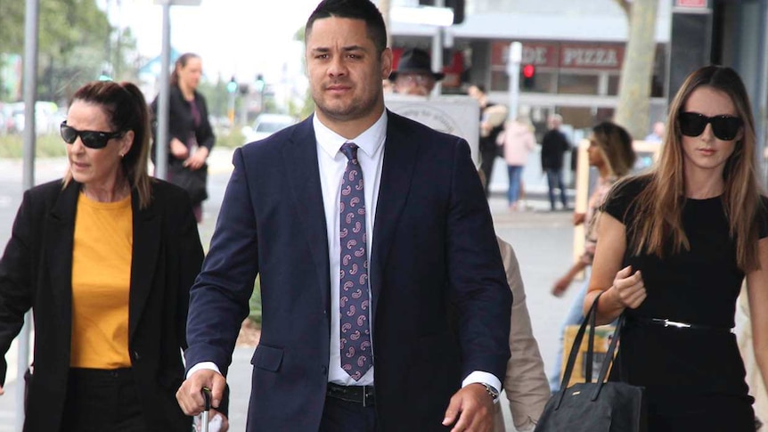 A man in a dark suit pulls a small suitcase down a city street, flanked by two women.