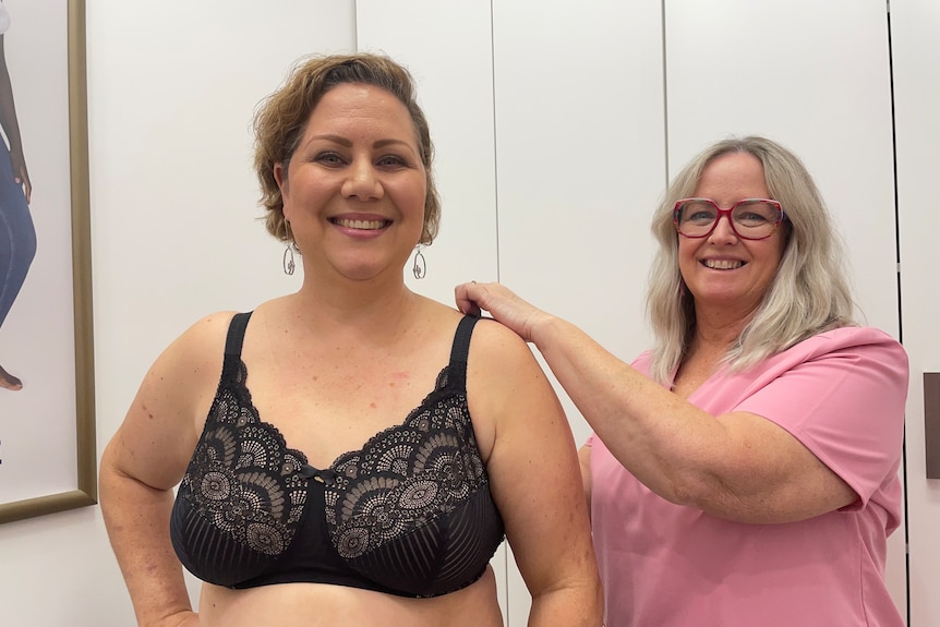 woman stands wearing bra while another woman with grey hair adjusting a bra strap 