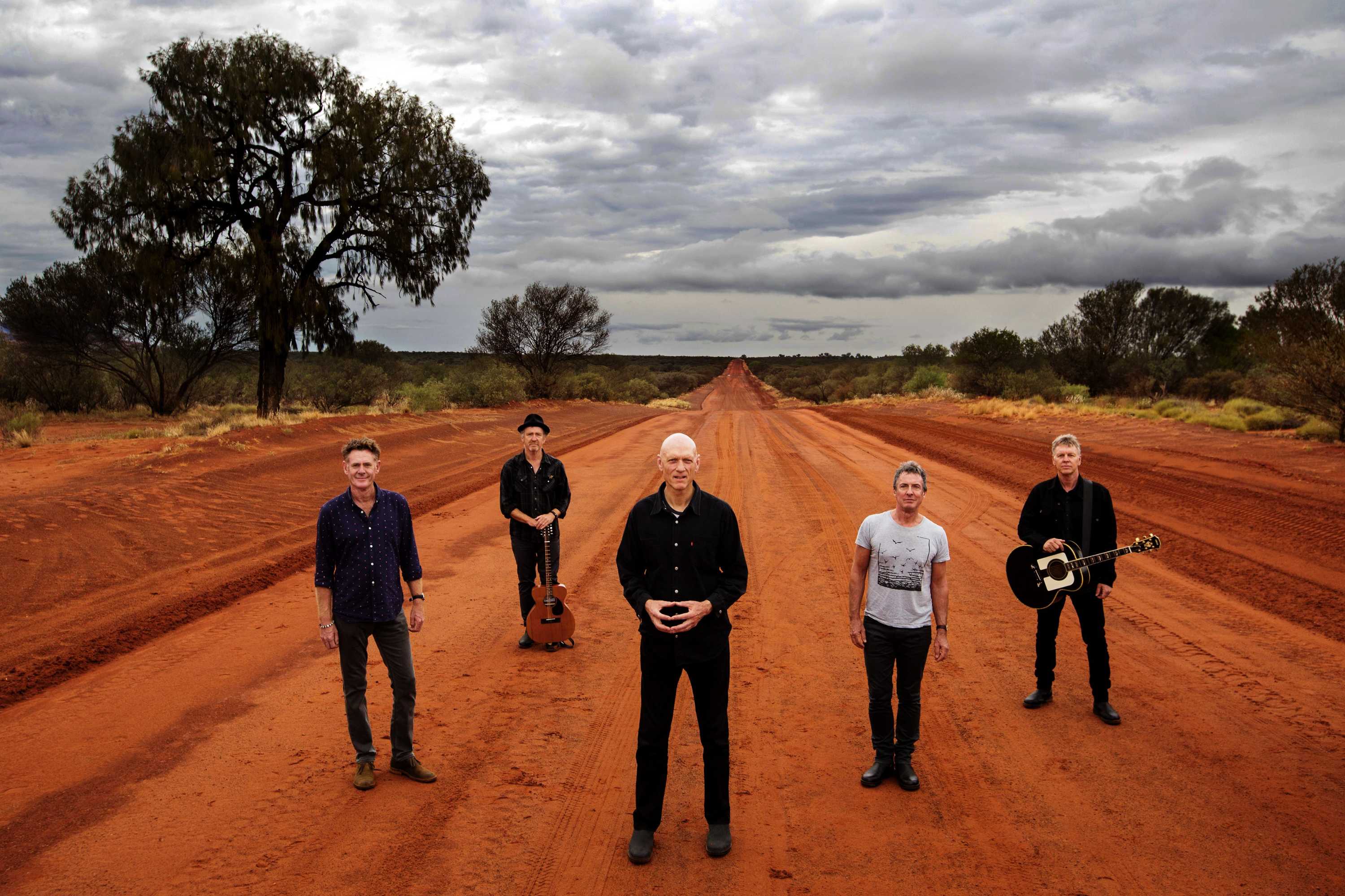 Five men (midnight oil) standing on a red dirt road. The road and shrubs to the side stretch to the horizon behind them