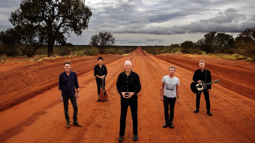 Five men (midnight oil) standing on a red dirt road. The road and shrubs to the side stretch to the horizon behind them