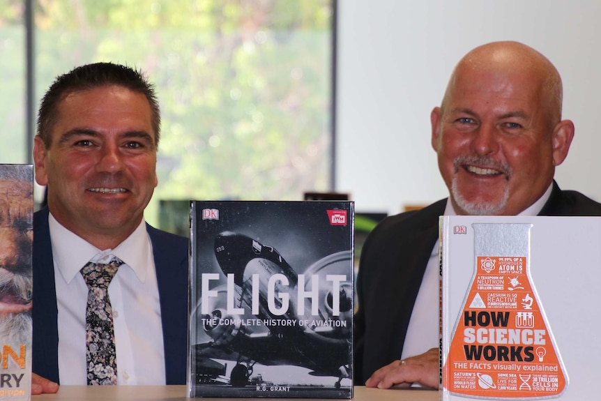 Two men sit smiling and posing for a photo with books in front of them.