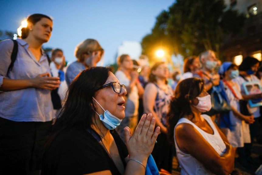 A woman with her hands clasped in front of her kneels as she is surrounded by others