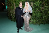 The designer John Galliano in his 60s in a black suit with Kim Kardashian in a silver dress and cardigan at the 2024 met gala
