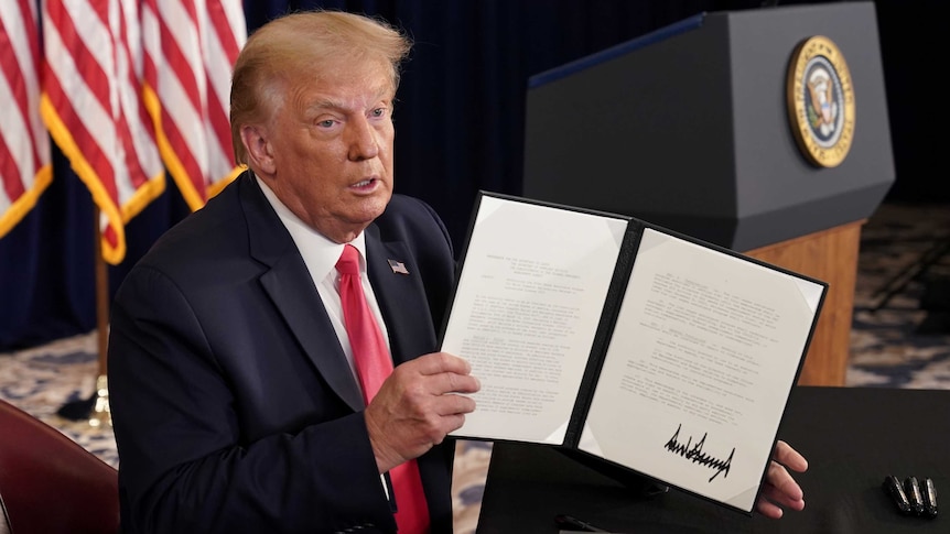 An elderly man in a dark suit holds a signed document in front of him with a flag and podium behind.