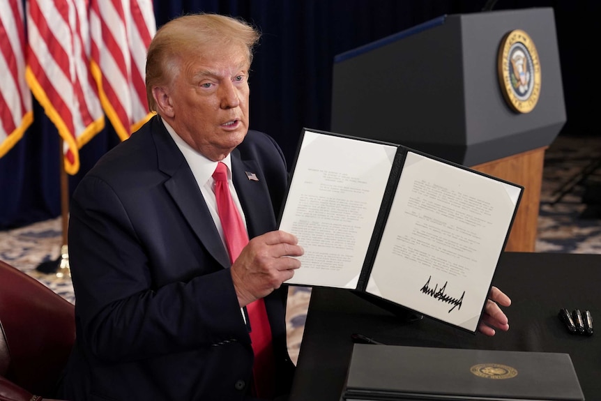 An elderly man in a dark suit holds a signed document in front of him with a flag and podium behind.