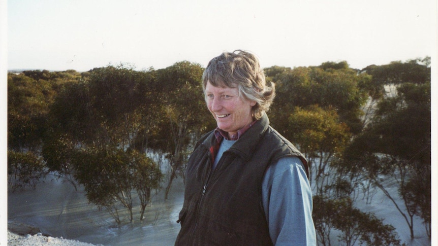 A woman standing on a sand dune with thin, small scrubby trees in the background.