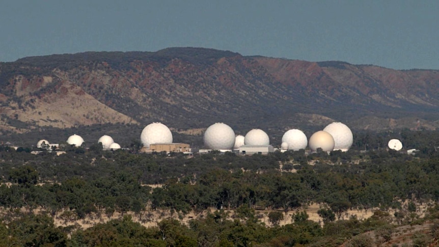 Large golf ball-like domes and low set buildings are surrounded by trees and bush. Behind them are hills.