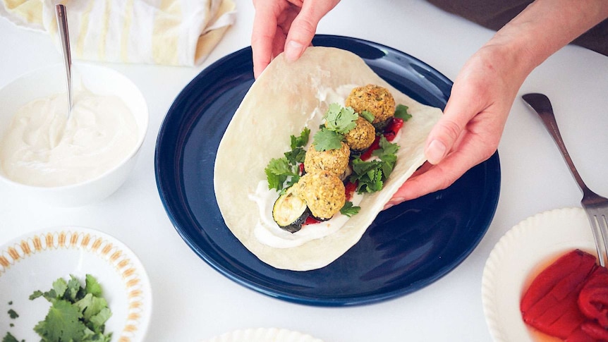 Four falafel balls sit atop a flatbread with yoghurt sauce, grilled capsicums, roasted zucchini and coriander, a family dinner.