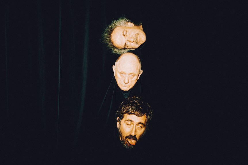 Three men stick their heads out of a black curtain and pull faces