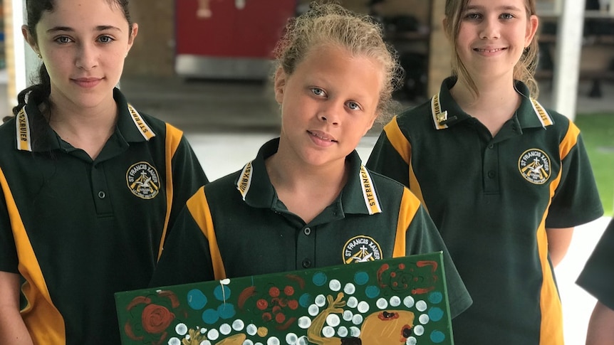 Two children in green and yellow in background, young boy with blond hair and blue eyes in foreground holding goanna painting.