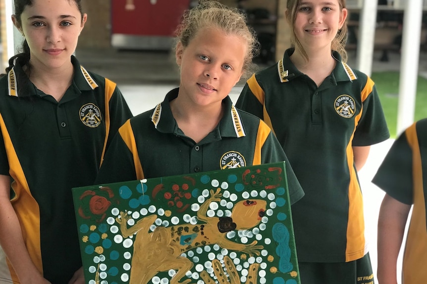 Two children in green and yellow in background, young boy with blond hair and blue eyes in foreground holding goanna painting.