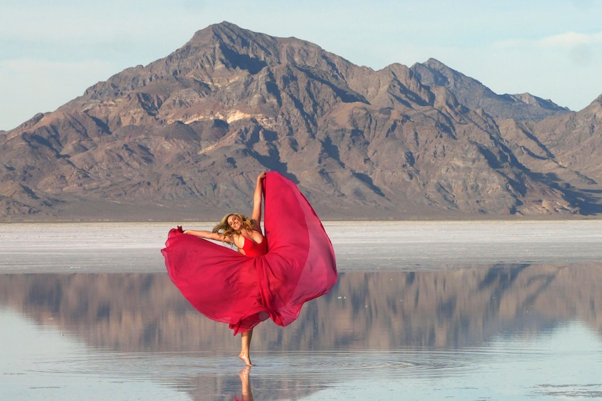 A woman holding the ends of a red dress up over a lake with a mountain in the background