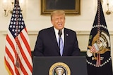 A still of Donald Trump speaking to a camera in front of a podium with two flags behind him.