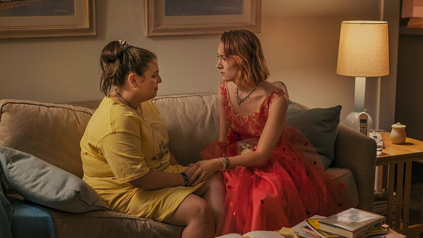 Colour still image of Saoirse Ronan and Beanie Feldstein having an emotional moment, sitting on a couch in a living room.