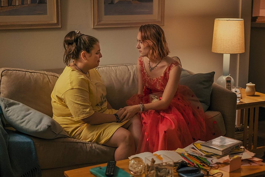 Colour still image of Saoirse Ronan and Beanie Feldstein having an emotional moment, sitting on a couch in a living room.