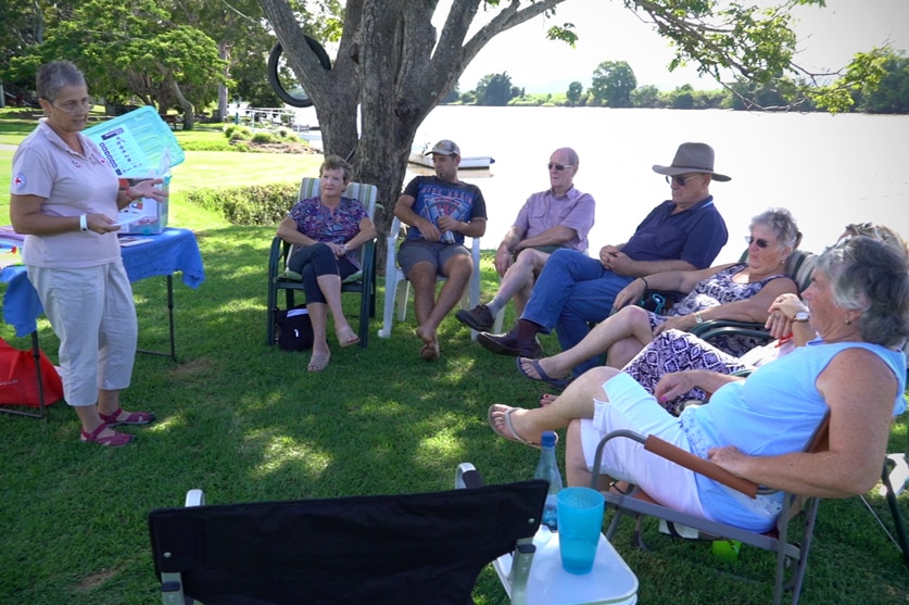 A woman instructs a group of seated people sitting on the banks of the Tweed River