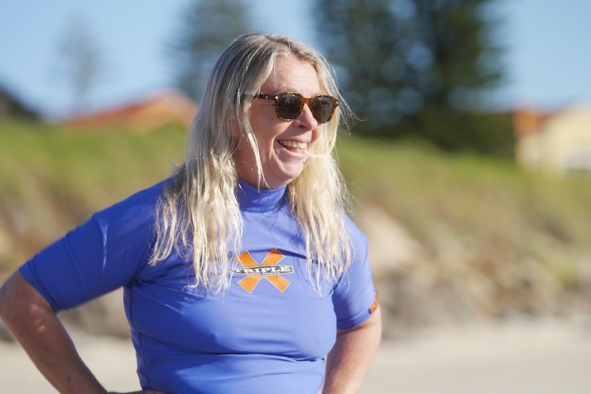 An older woman with long blonde hair and sunglasses smiles. She wears a blue rash top and the beach behind her is out of focus.