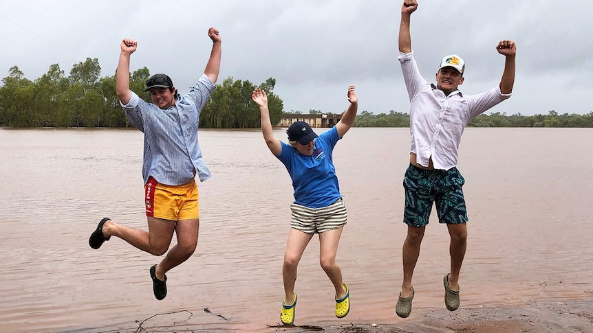 Three residents jump in the air with joy next to floodwaters for the rain at drought-stricken Cloncurry.