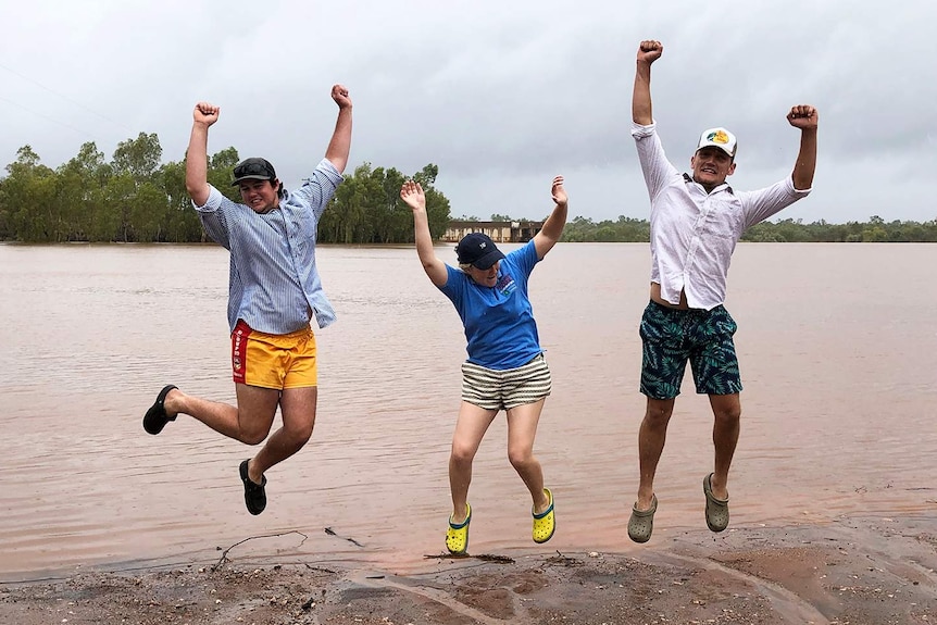 Three residents jump in the air with joy next to floodwaters for the rain at drought-stricken Cloncurry.