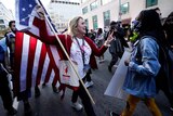 a woman holding a United States flag holds up two fingers as she approaches a woman in a mask