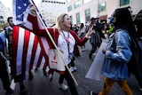 a woman holding a United States flag holds up two fingers as she approaches a woman in a mask
