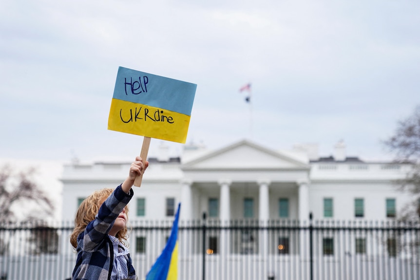 A sign that reads 'Help Ukraine' is held up by a child in the foreground, behind them is The White House in Washington DC.