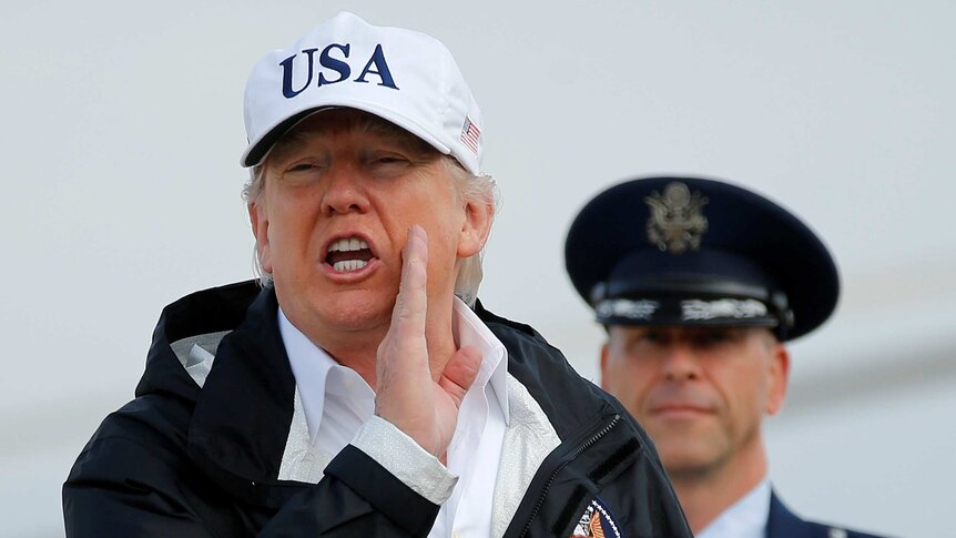Donald Trump answers a reporter's question wearing a USA hat and holding his right hand to his left cheek.
