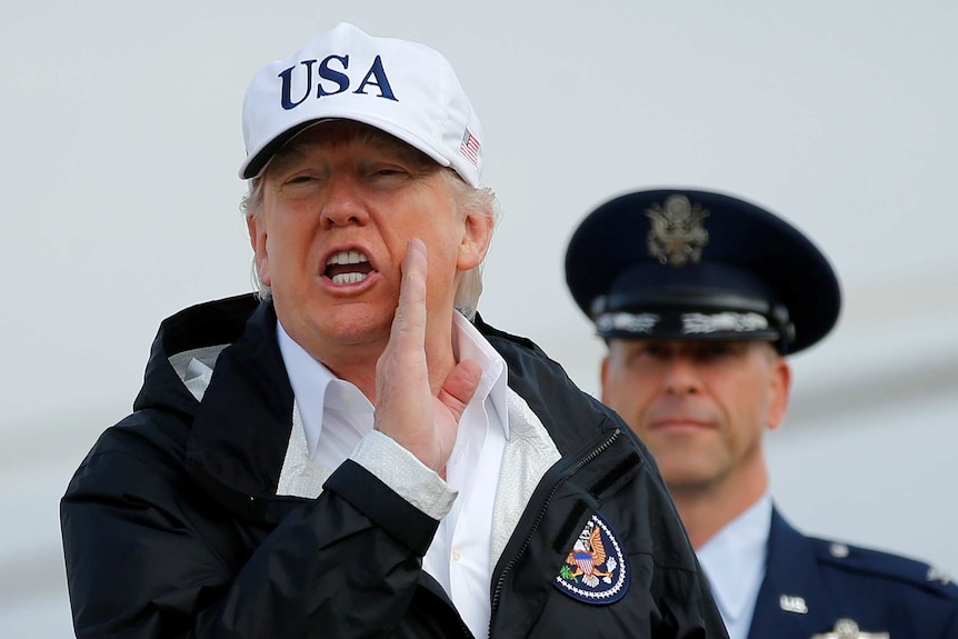 Donald Trump answers a reporter's question wearing a USA hat and holding his right hand to his left cheek.