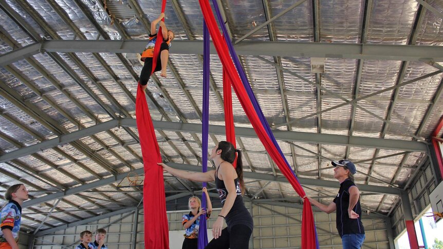 A young girl holds on to a ribbon suspended from the roof of a sports hall, a teacher and class mates watch on.