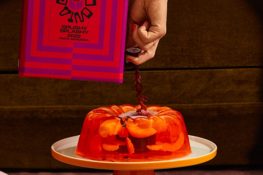 Hellhäutige Hand, rote Nägel, gießt vor einer dunklen Wand Rotwein aus einem rot-rosa gestreiften Fass auf Orangenfruchtgelee-Kuchen.