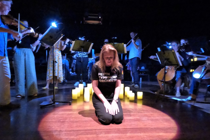 A woman wears a Perth Symphony Orchestra t-shirt as she sits in the centre of a spotlight surrounded by musicians