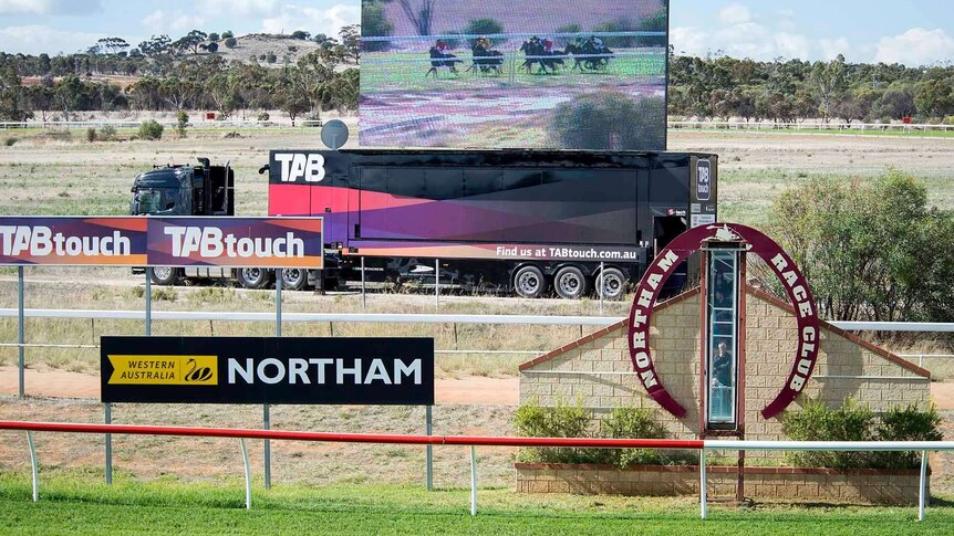 A big screen standing on the back of a semi-trailer behind the winning post at Northam Race Club shows a horse race in progress.