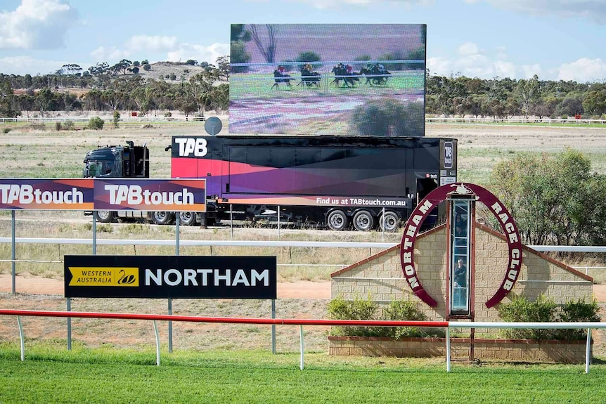 A big screen standing on the back of a semi-trailer behind the winning post at Northam Race Club shows a horse race in progress.