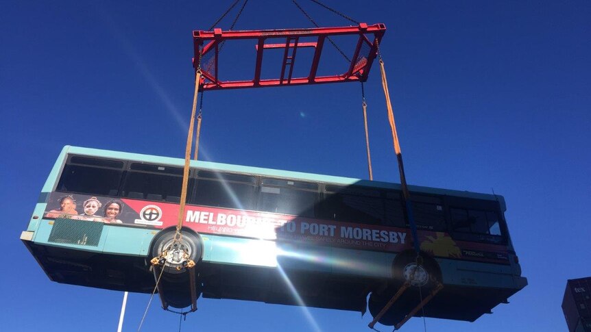 A blue bus bearing a sign Melbourne to Port Moresby is unloaded off a ship in Port Moresby by crane