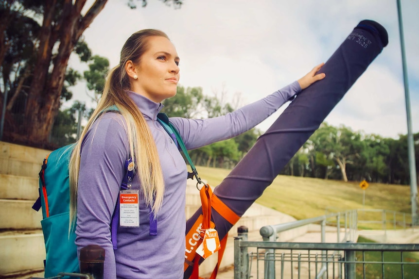 A caucasian woman dressed in a long-sleeve purple jacket and a blue backpack looks out onto the field with her javelin.