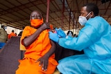 An African man in an orange jumpsuit and mask receives a vaccination from an African man in a mask and blue protective suit