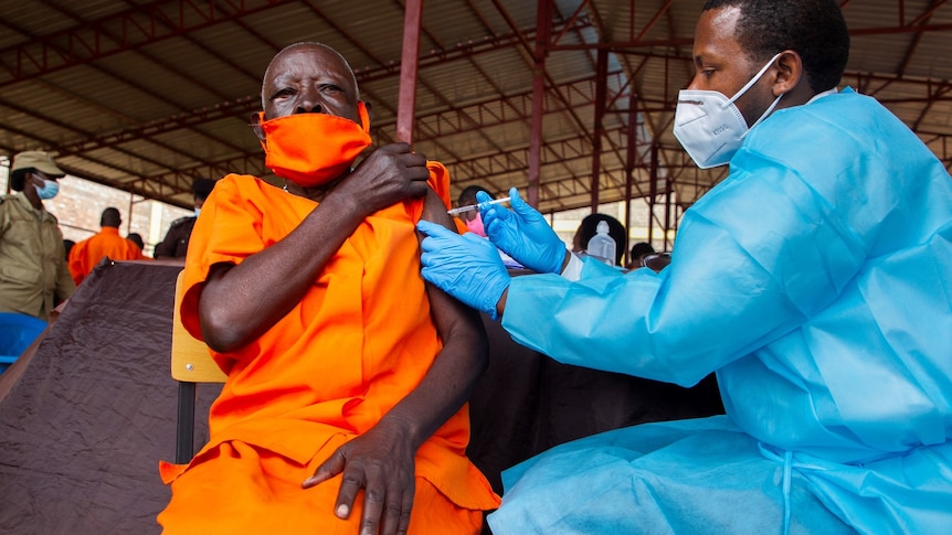 An African man in an orange jumpsuit and mask receives a vaccination from an African man in a mask and blue protective suit