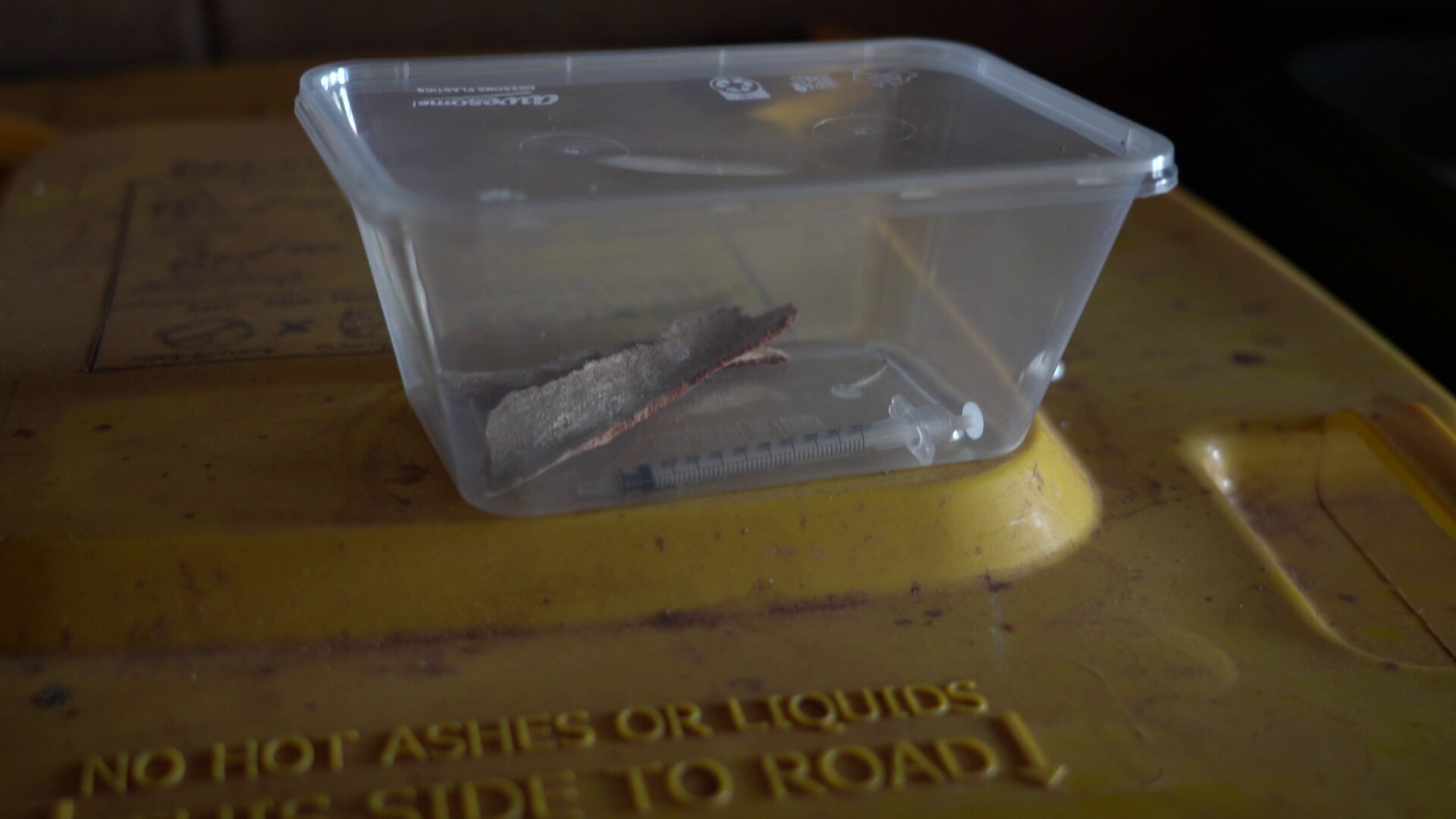 Una aguja abierta en un contenedor transparente de lonchera para llevar sobre un bote de basura. 