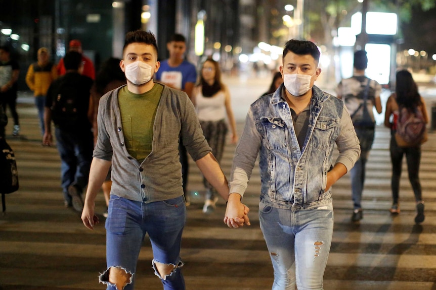 Two men in face masks hold hands crossing a street at night