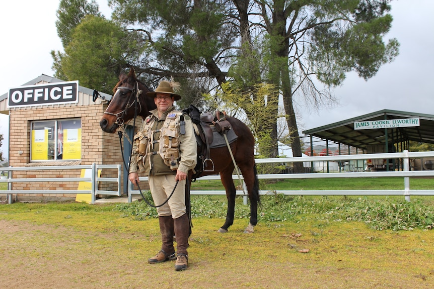 A man dressed in Army regalia stands next to a bay hose in a paddock.