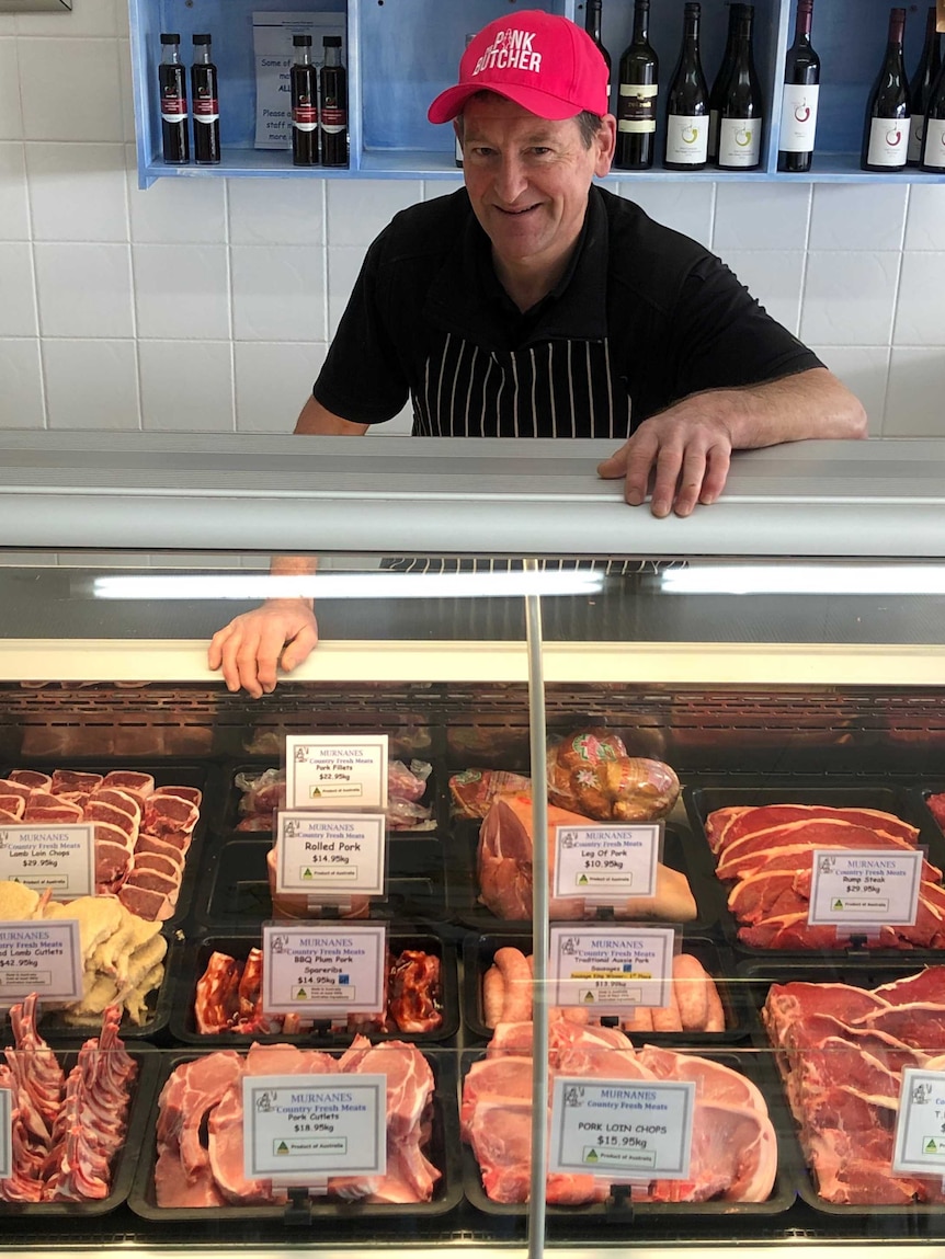 Butcher in his shop wearing a jaunty red cap and smiling over his chops.